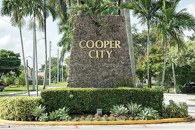 Stone monument sign reading Cooper City surrounded by palm trees and manicured green foliage.